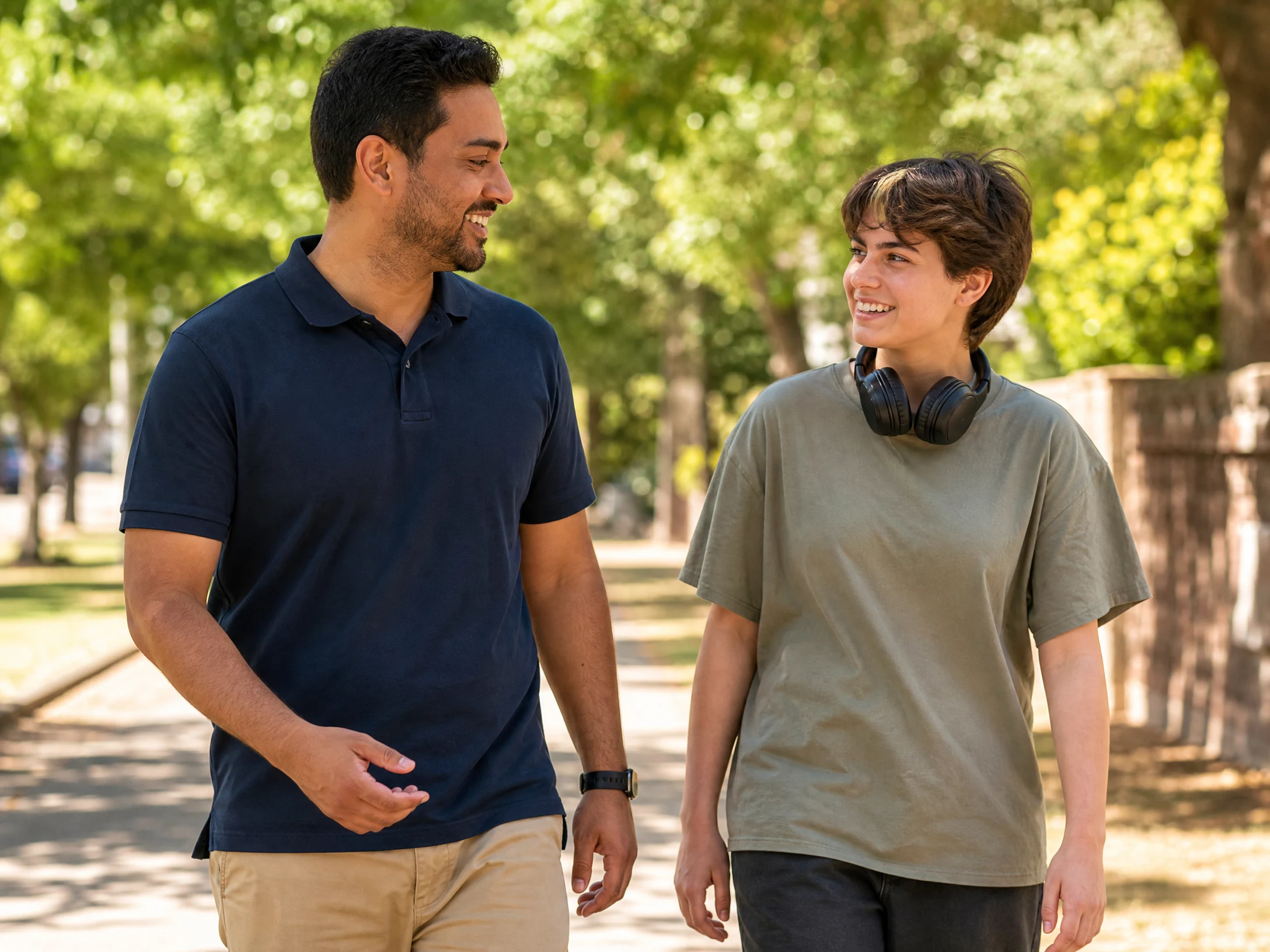 Support worker in navy polo shirt walking and talking with a young participant along a sunny tree-lined path