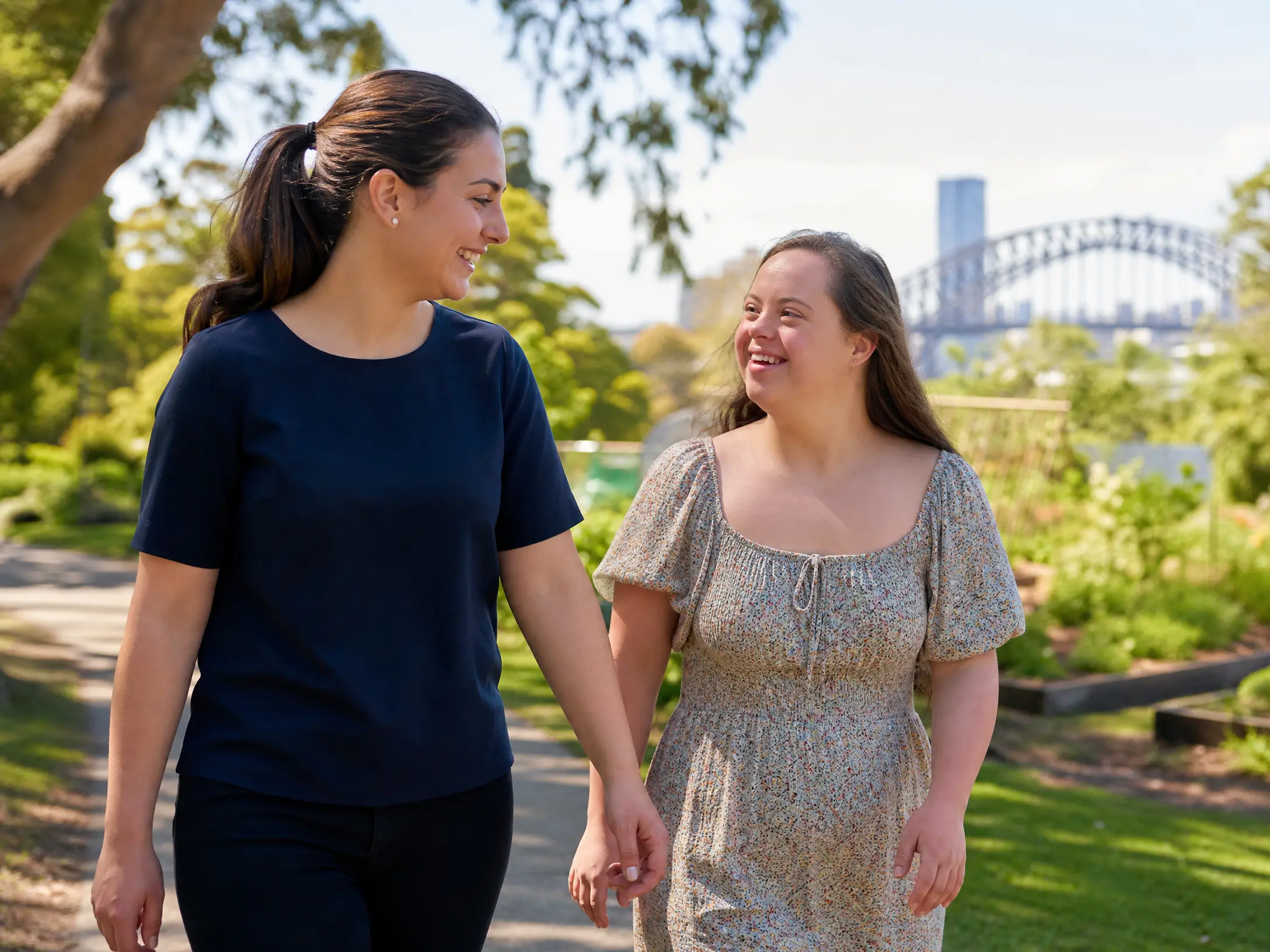 A Lanara support worker in a navy top walking hand-in-hand with a young female participant with Down syndrome through a park near Sydney Harbour Bridge, both smiling warmly