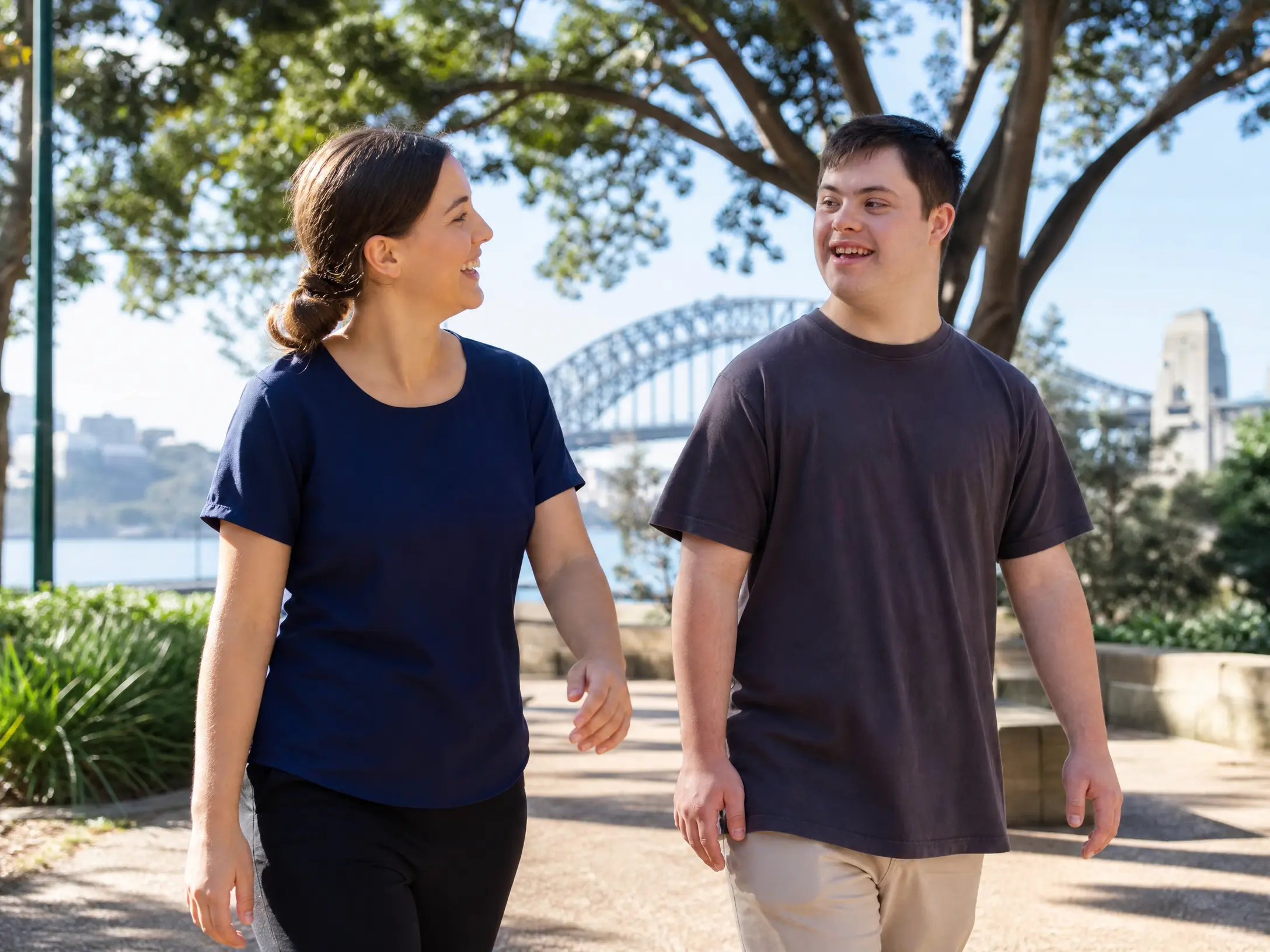 A Lanara support worker in a navy top walking and talking with a young male participant in a Sydney park with the Harbour Bridge visible in the background